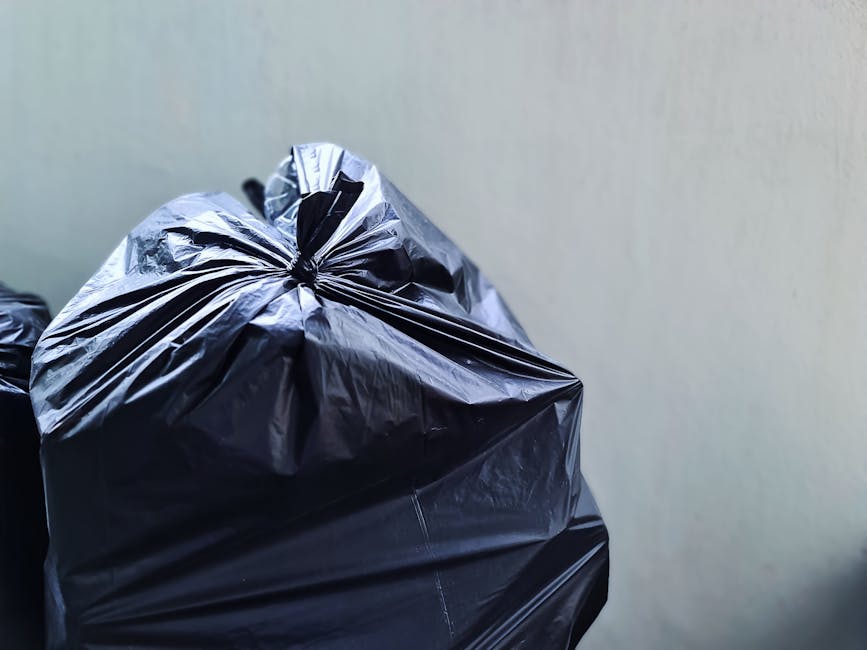 A close-up of multiple black plastic rubbish bags tightly knotted at the top, positioned in the foreground against a plain, light-colored wall. The bags are made of glossy, thick plastic material, ref