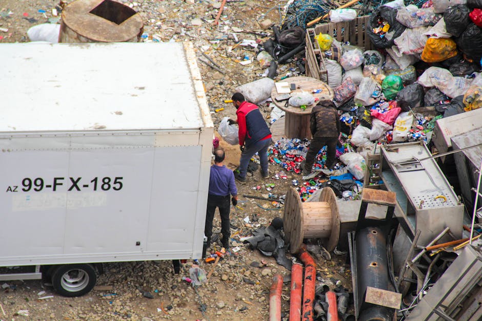 A large, irregular pile of mixed domestic rubbish and waste materials scattered across a paved outdoor area. The collection includes crumpled paper, plastic bottles, glass jars, and metal containers, 